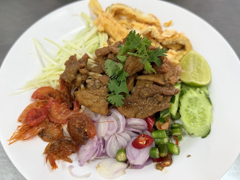 ข้าวคลุกกะปิในจานขาว มีหมูหวาน ไข่ฝอย กุ้งแห้งทอด มะม่วงซอย หอมแดง พริก แตงกวา และมะนาว