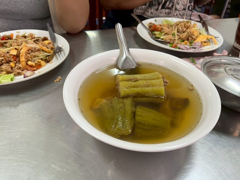 ต้มมะระยัดไส้ในชามขาว น้ำซุปใส มีจานข้าวคลุกกะปิวางอยู่ด้านหลัง