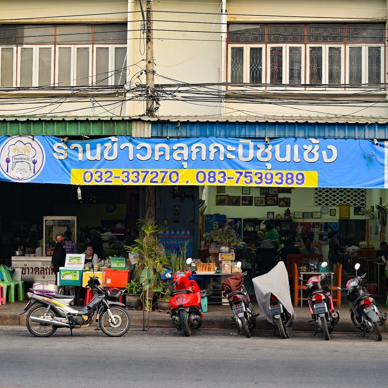 หน้าร้านมองจากฝั่งตรงข้ามถนน ป้ายสีน้ำเงินยาวตลอดสองคูหา